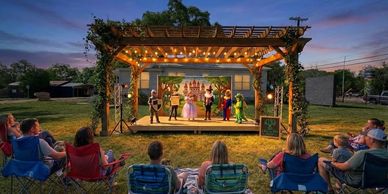 Outdoor theater performance at sunset with audience seated on lawn chairs.