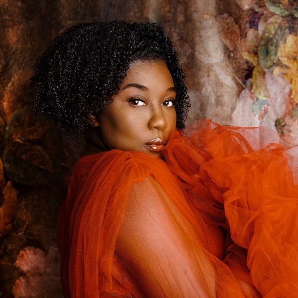 A woman in an orange sheer dress poses against a textured background.