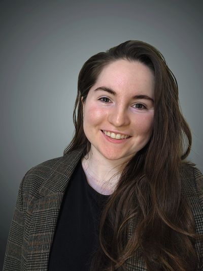 Smiling young woman with long brown hair wearing a checkered blazer and black top.