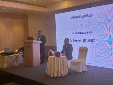 Speaker delivering keynote address at a conference stage with a seated guest.