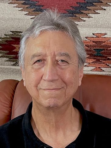 Casual portrait of George Glaser sitting in a chair with American Indian rug on the wall behind.