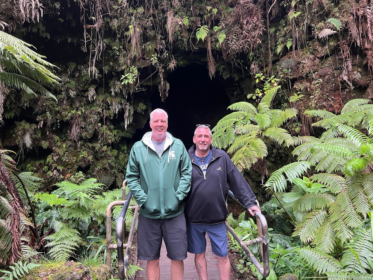 Two men standing on a bridge in front of a dark cave surrounded by lush green ferns.