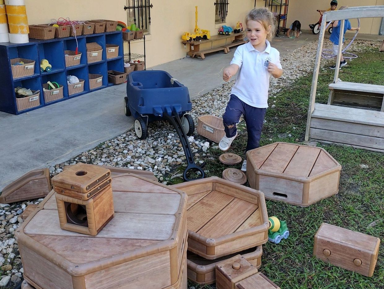 Young child playing outdoors with wooden educational toys and a blue wagon.