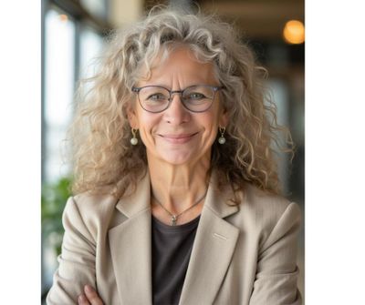 Smiling woman with curly gray hair and glasses in a beige blazer.