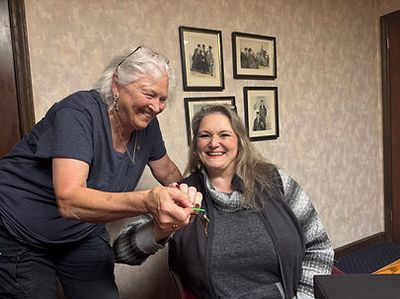 Two women smiling and holding hands indoors with framed photos on the wall.