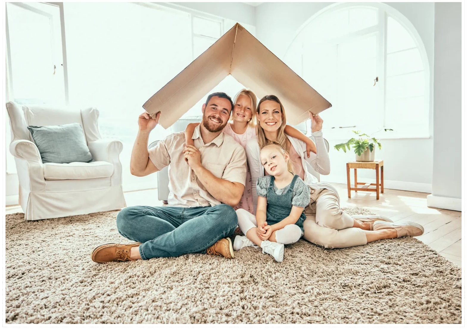 A family of four sitting in their living room smiling holding a piece of cardboard over their head 