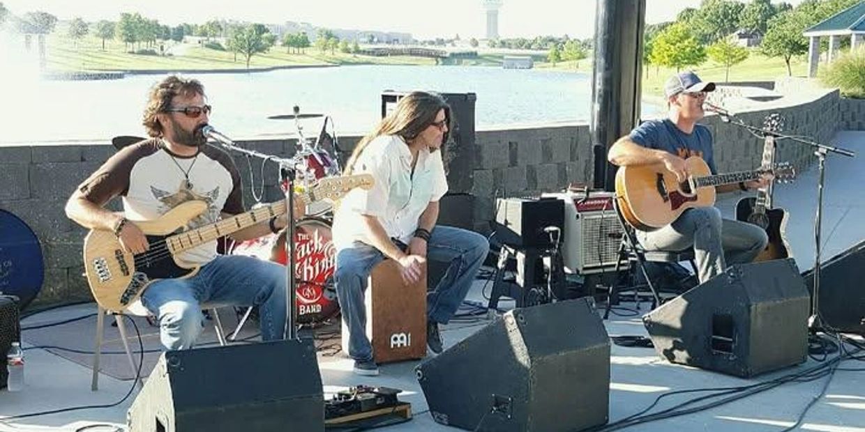Three musicians performing outdoors by the water with guitars and percussion.