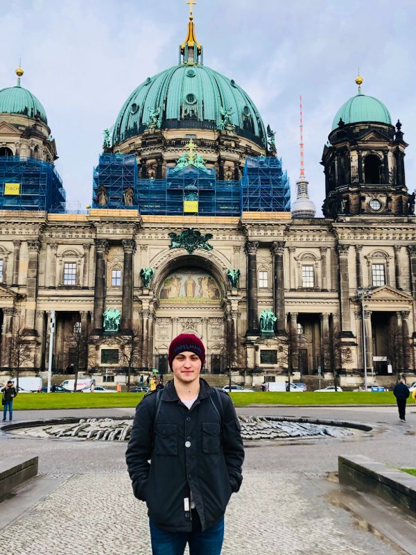 A young man stands in front of a historic cathedral with green domes under a cloudy sky.