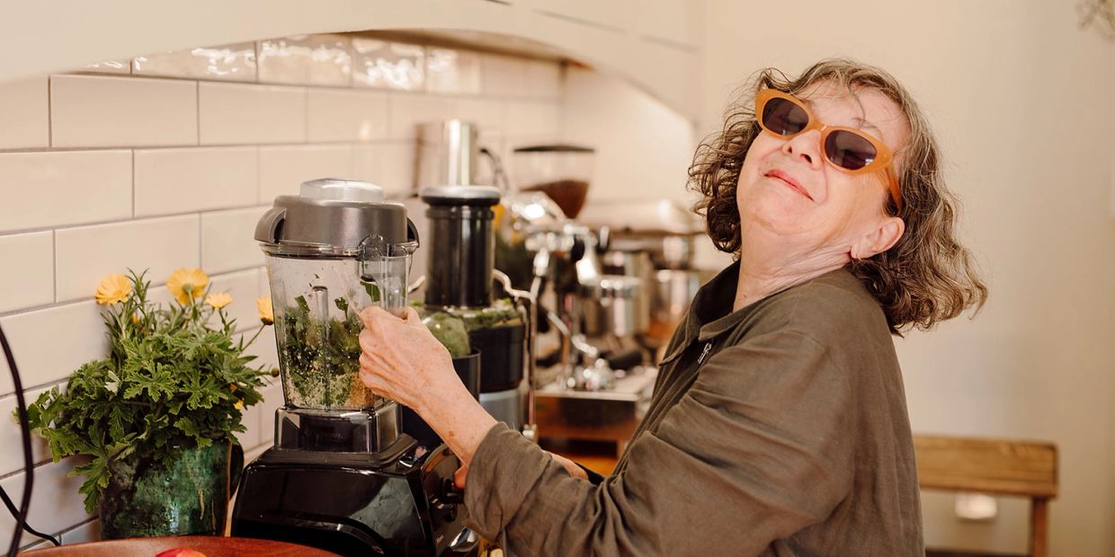 Woman blending greens in kitchen with sunglasses.