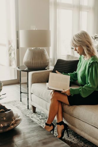 Woman in green blouse reading a book on a beige couch in a bright living room.