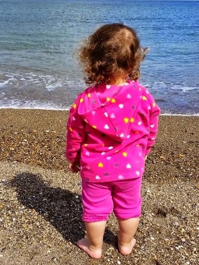 A toddler in pink clothes stands barefoot on a pebble beach facing the sea.