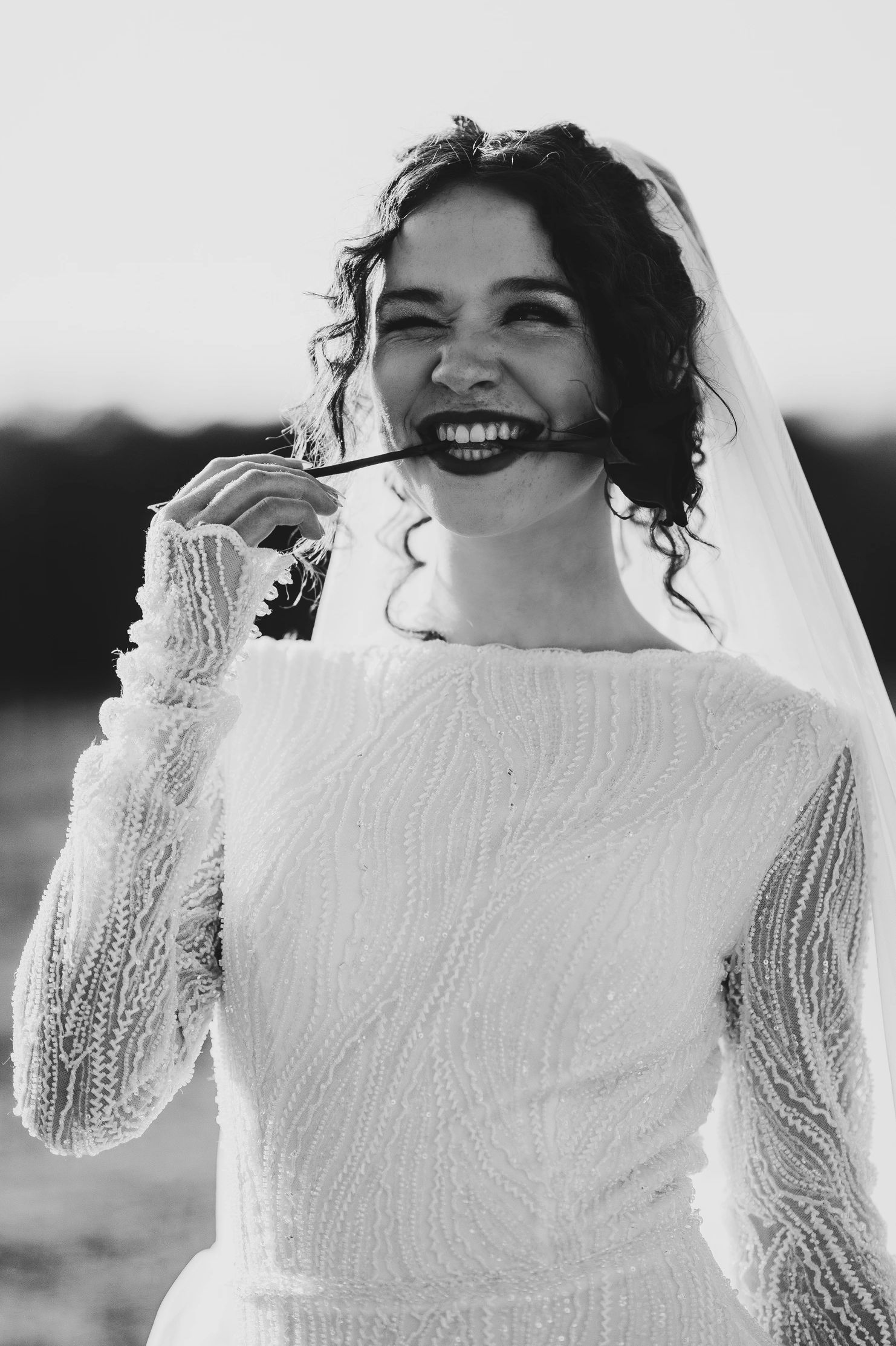 Black-and-white portrait of a joyful bride, smiling with a flower stem in her teeth; veil glows in s