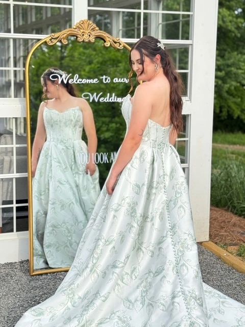 Bride in a pale green floral jacquard strapless ball gown admiring her reflection in a vintage gold mirror with “Welcome to our wedding” written on it.