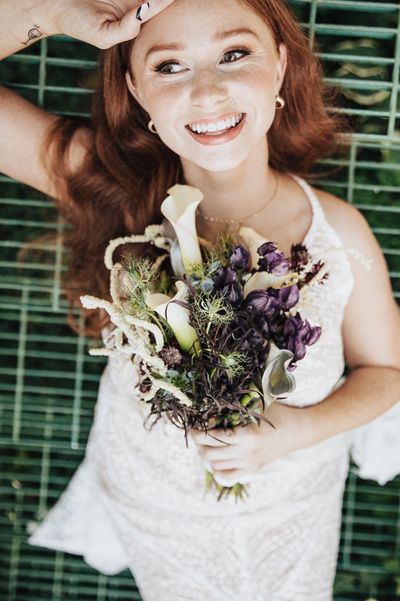 Smiling bride holding a purple and white bouquet, lying on a green metal grate.