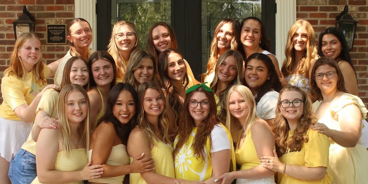 Group of young women smiling together in yellow and white outfits outside a brick building.