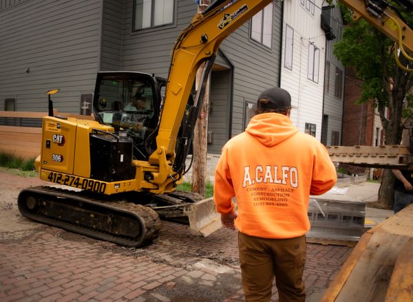 A worker in an orange hoodie supervises a yellow excavator at a construction site.