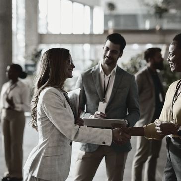 Two professional women shaking hands at a business event with others networking in the background.