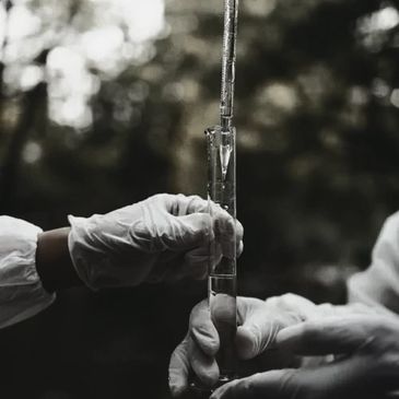 Hands in gloves handling a graduated cylinder outdoors.