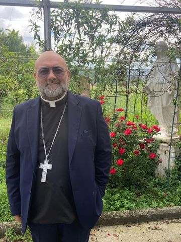 Clergyman with a cross necklace stands near a garden with red flowers and a statue.