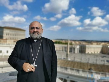 A clergyman wearing a black robe and cross necklace stands outdoors with a cityscape background.