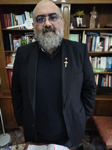Bearded man wearing glasses and a black suit with a cross pin, standing in front of a bookshelf.