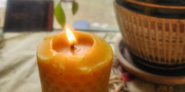 Lit yellow candle in focus with a plant pot in the background.