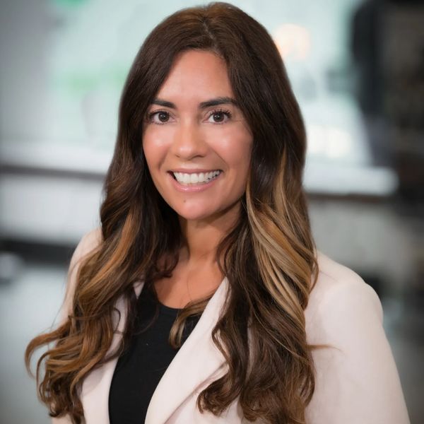 A smiling woman with long wavy brown hair wearing a light blazer.