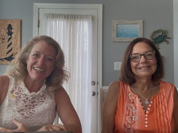 Two smiling women sitting indoors with decorative wall art behind them.