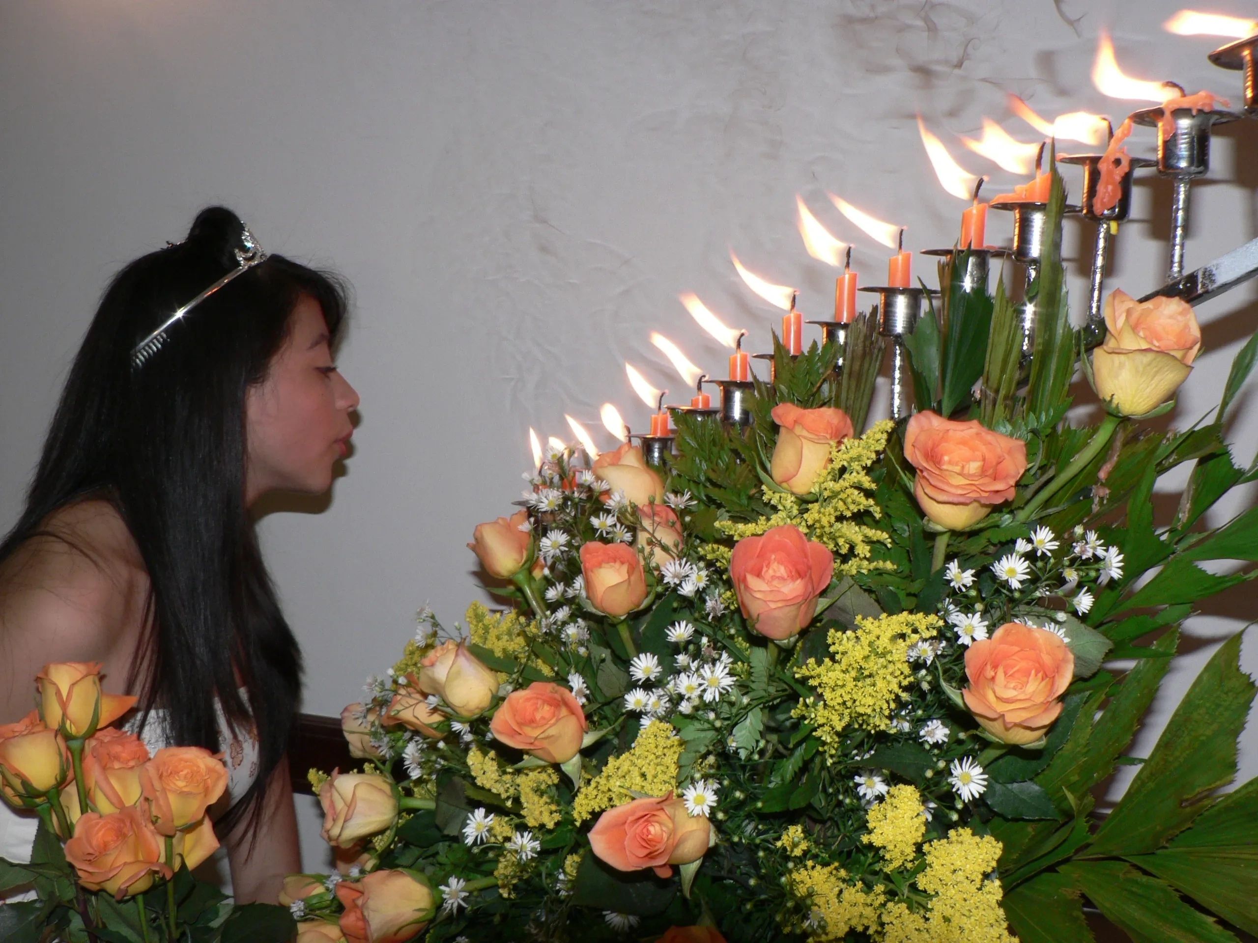 Young woman with tiara lighting candles amidst orange roses and greenery.