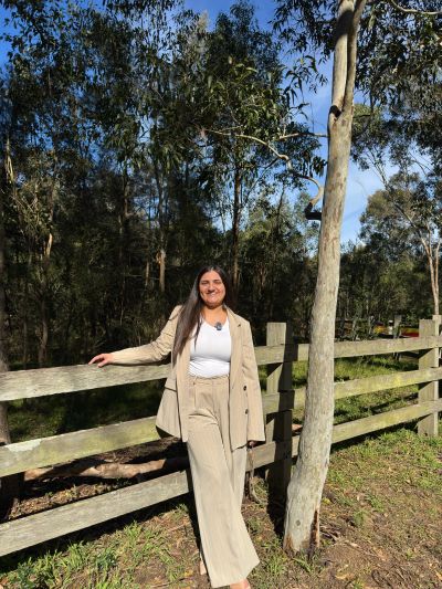 Woman dressed in beige leaning on wooden fence outdoors.