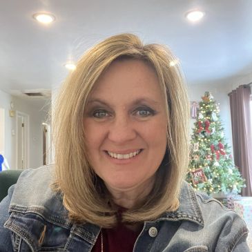 Smiling woman with blonde hair in a denim jacket, Christmas tree in background.
