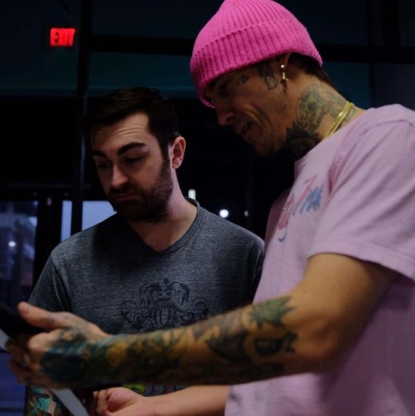 Two men examining a tattoo design, one wearing a pink beanie and shirt with tattoos.