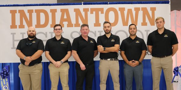 Six men in black shirts and khakis pose in front of an Indiantown High School banner.