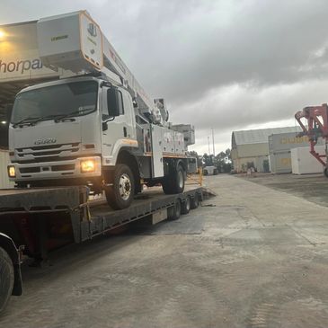 Isuzu truck with crane loading on a flatbed trailer in an industrial area.