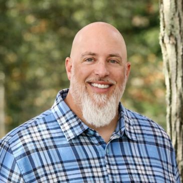 Smiling bald man with a white beard wearing a blue plaid shirt outdoors.