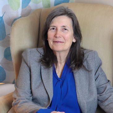 A woman in a blue blouse and gray blazer sits in a beige armchair.