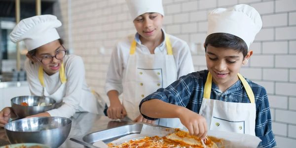 Three kids wearing chef hats and aprons preparing pizza together.
