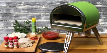 A green portable pizza oven with ingredients and a pizza peel on a wooden table.