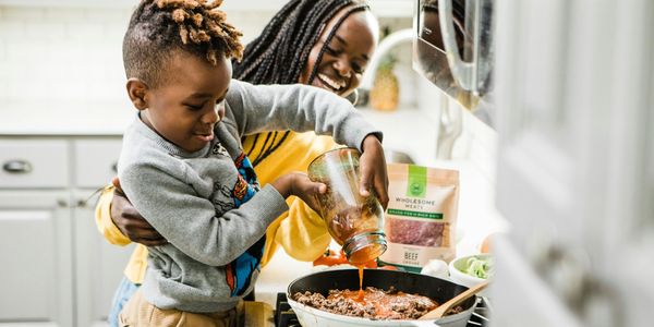 A child joyfully helps pour sauce into a pan while an adult watches in a kitchen.