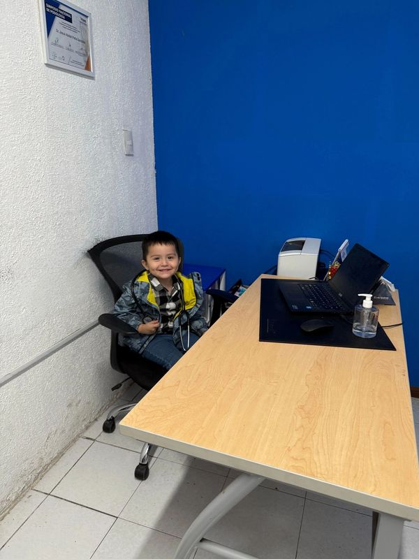 A young boy smiling while sitting at an office desk with a laptop and hand sanitizer.