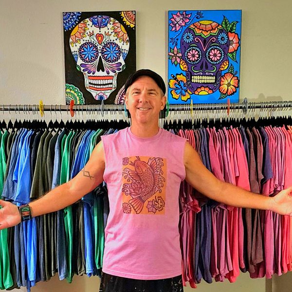 Man in pink sleeveless shirt stands smiling in front of colorful hanging clothes and vibrant skull paintings.