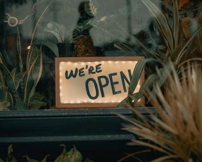 Lit sign saying "We're Open" in a shop window surrounded by plants.