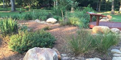 A peaceful garden with rocks, plants, and a wooden bridge in sunlight.