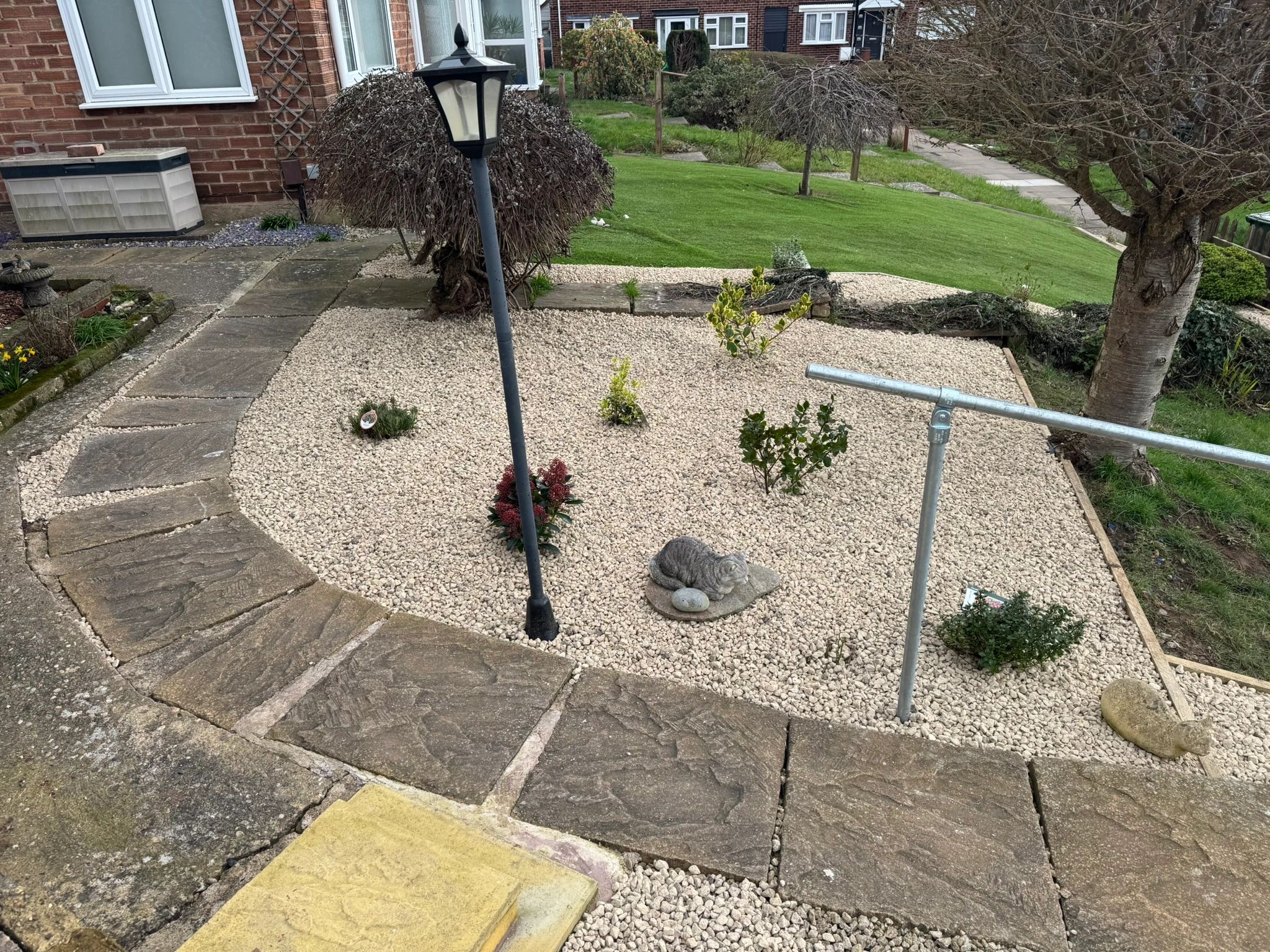 Stone pathway leading to a garden with shrubs, a lamp post, and a cat statue.