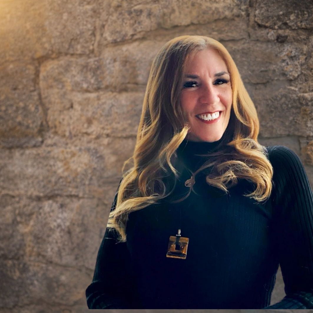 Smiling woman with long blonde hair against a stone wall background.