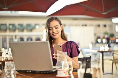 Woman looking at phone and laptop at outdoor cafe.