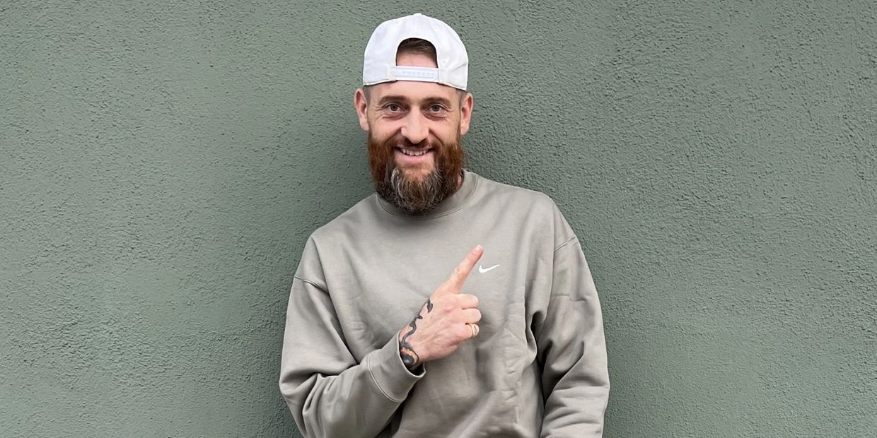 Bearded man smiling and pointing up, wearing a white cap and gray sweatshirt.