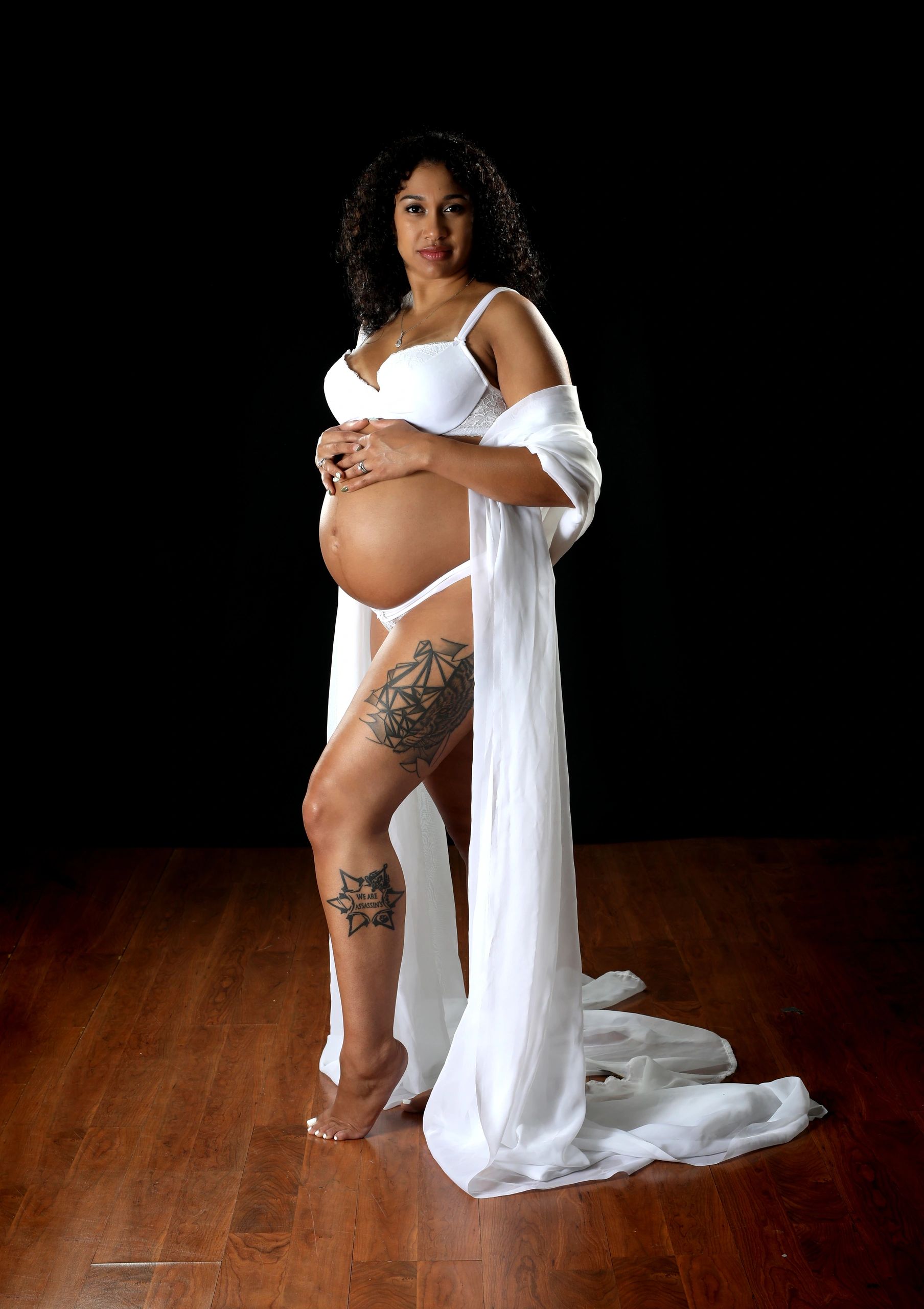 Pregnant woman in white lingerie and sheer robe on wooden floor.