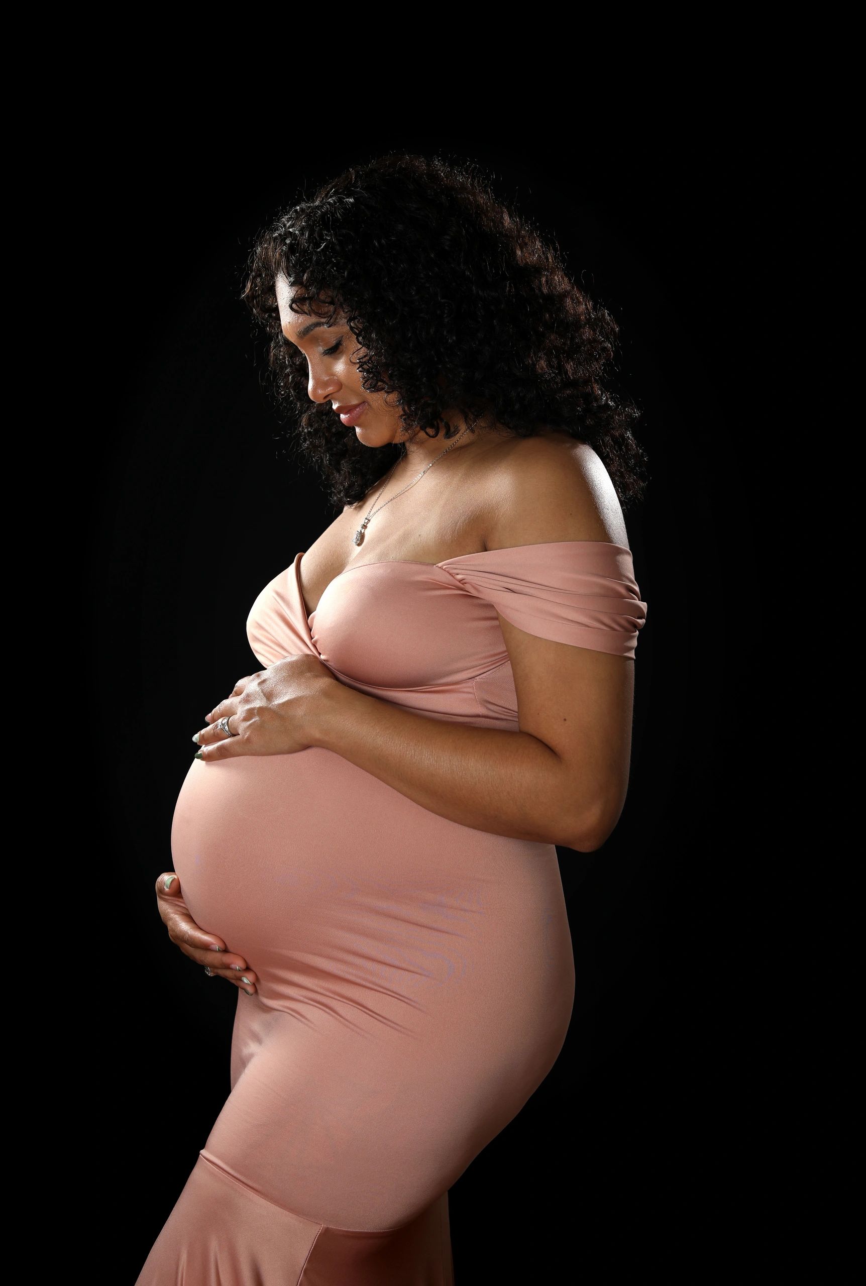 Pregnant woman in a pink dress gently holding her belly against a black background.