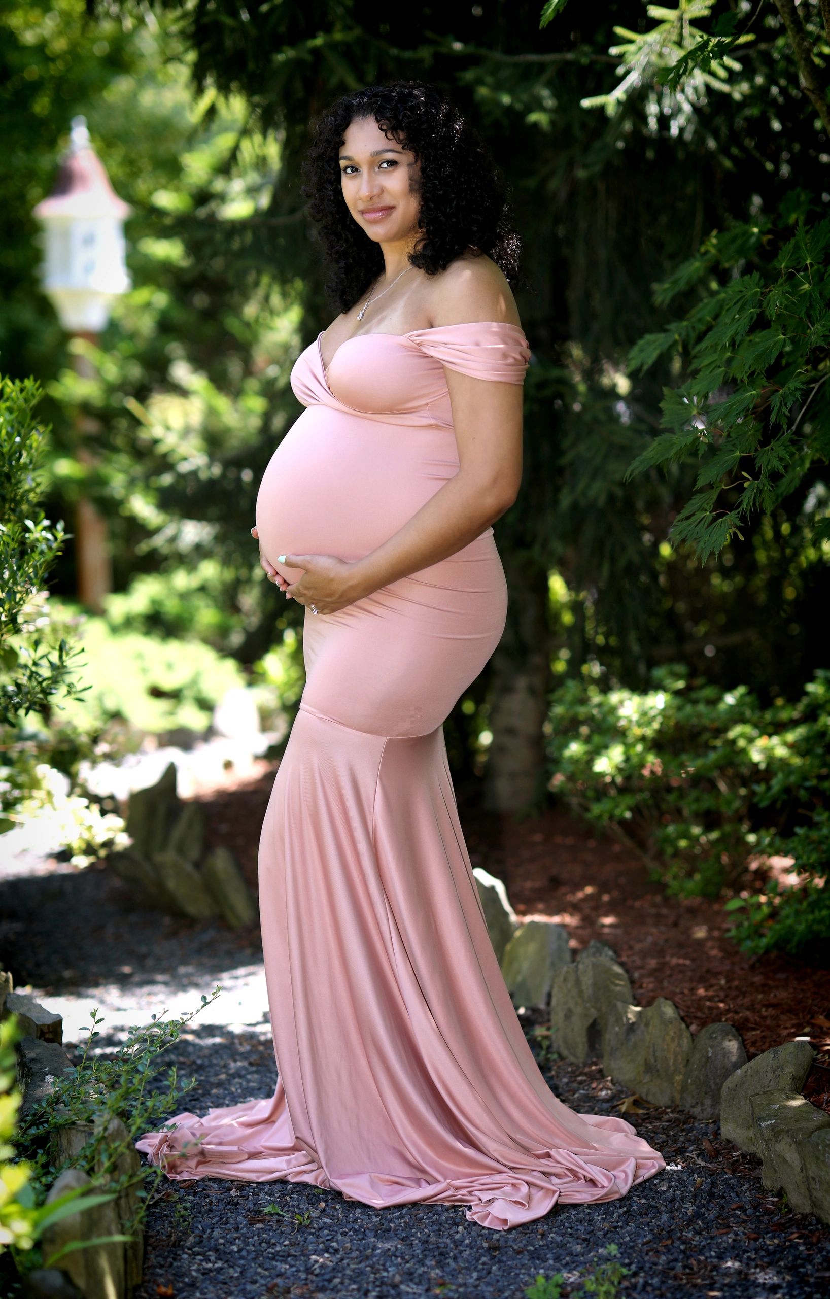 Pregnant woman in a flowing pink dress standing outdoors surrounded by greenery.
